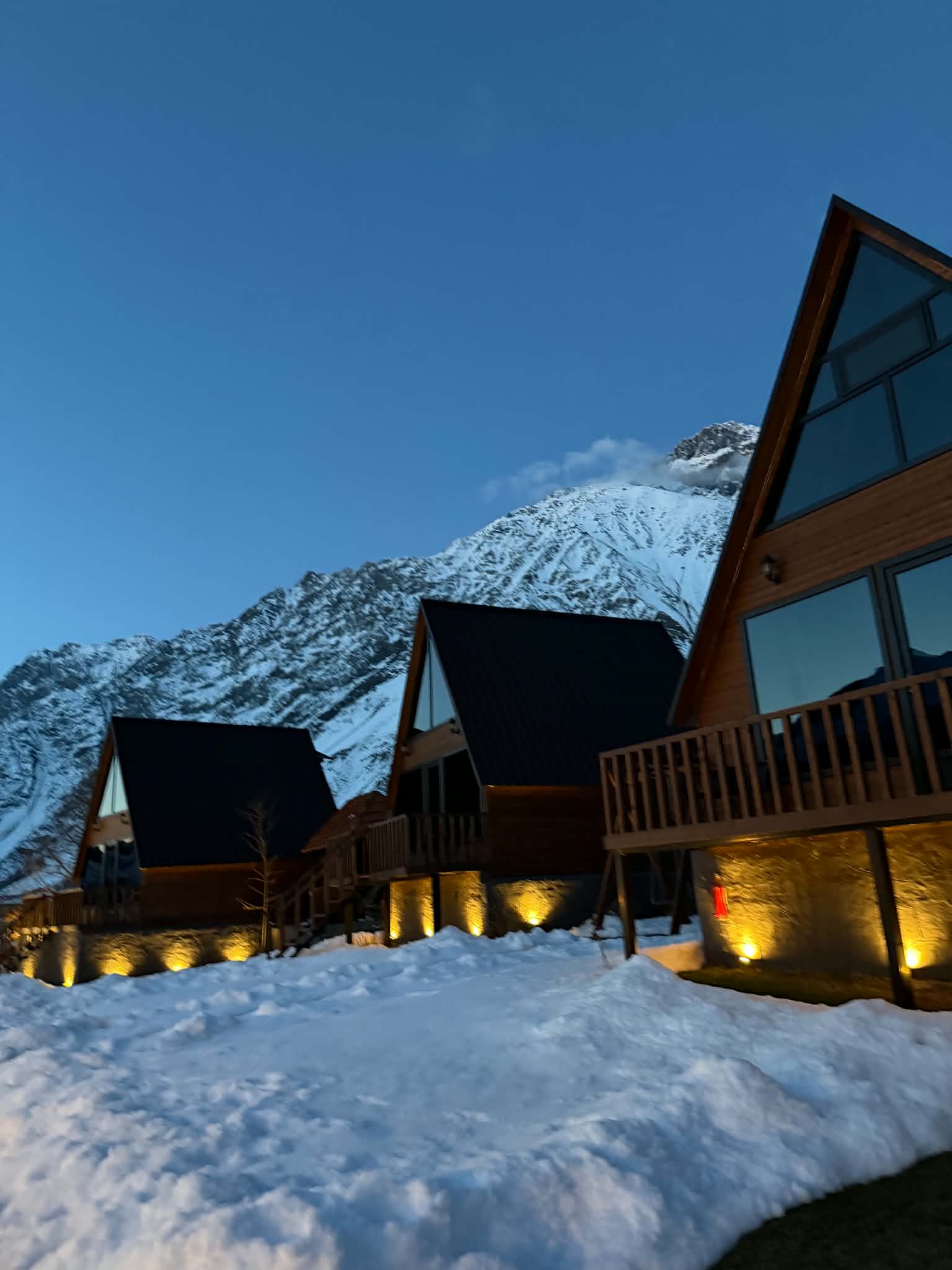 Three A-frame wooden cottages glowing at dusk beneath snow-capped Mt. Kazbek